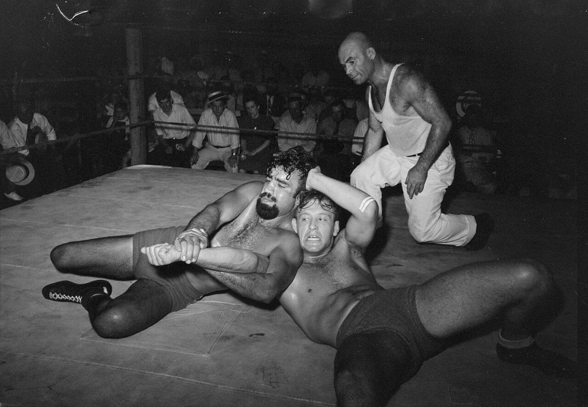 A wrestling match performance in Sikeston, Missouri, May 1938. Two wrestlers grapple while the referee (in white, right) looks on. The beard